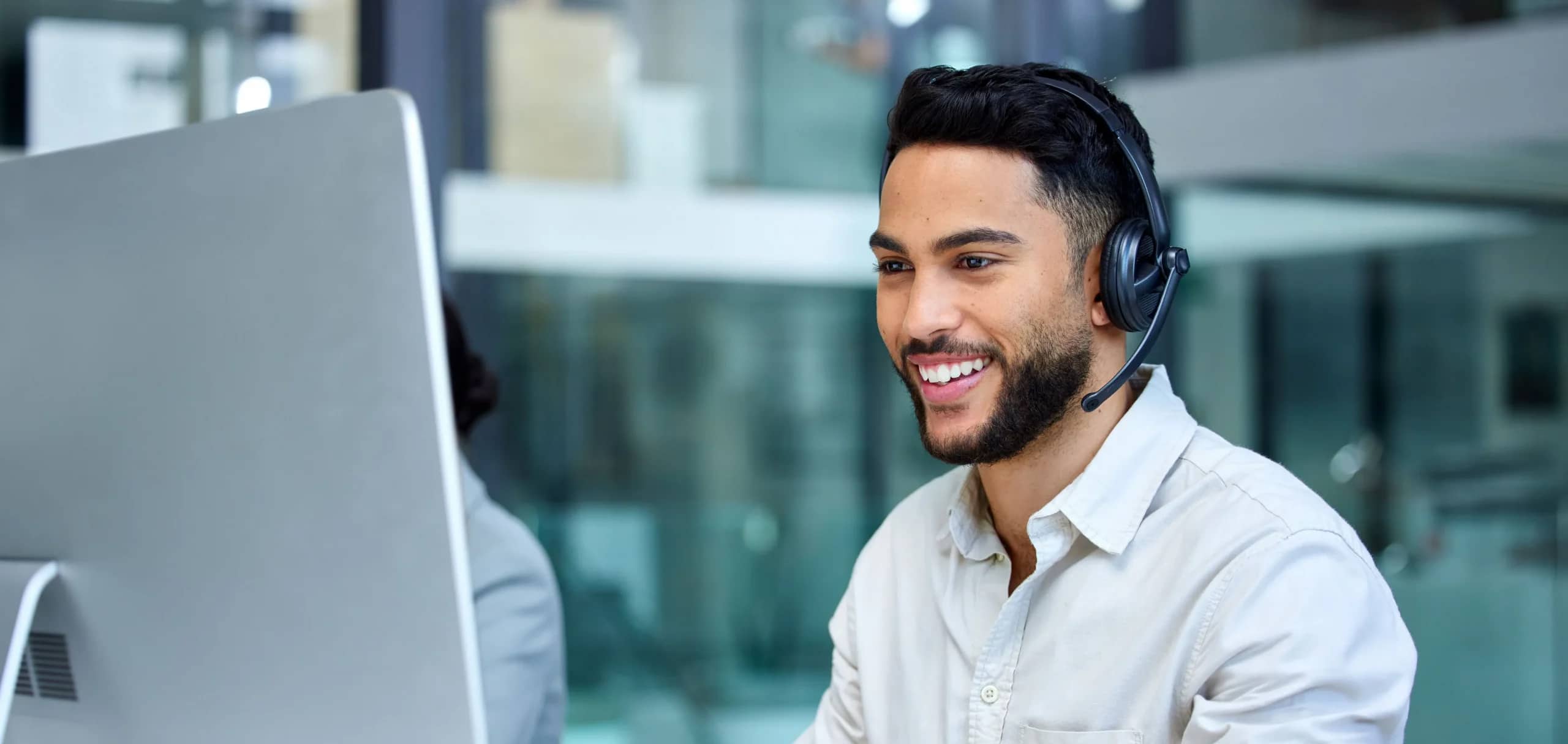 A smiling man with a beard wearing a headset sits at a desk, looking at a computer monitor in a modern office environment.