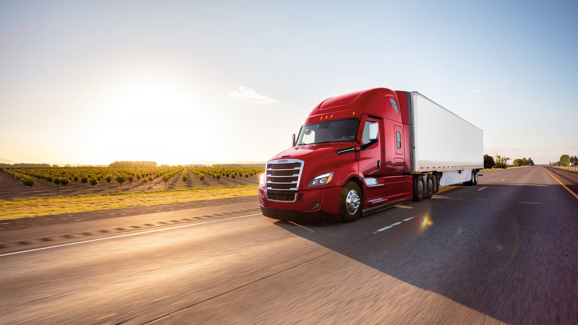 A red semi-truck with a white trailer drives down a highway at sunrise or sunset, passing farmland and open fields under a clear sky.